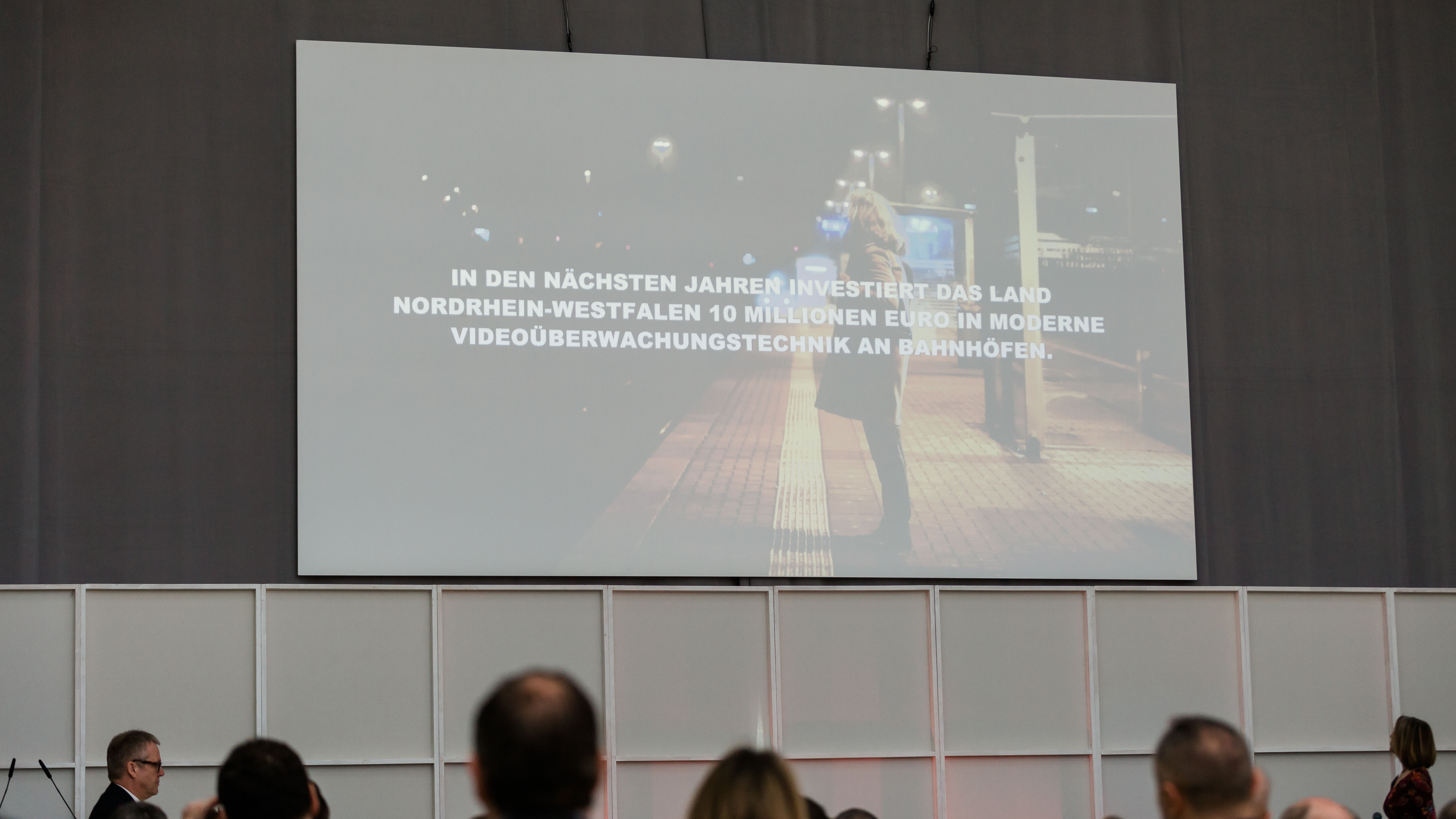 Eine Folie der Präsentation auf einer großen Leinwand. Dort zu lesen: "In den nächsten jahren investiert das Land NRW 10 Millionen Euro in moderne Videoüberwachungstechnik an Bahnhöfen". - öffnet Bild in größerer Ansicht