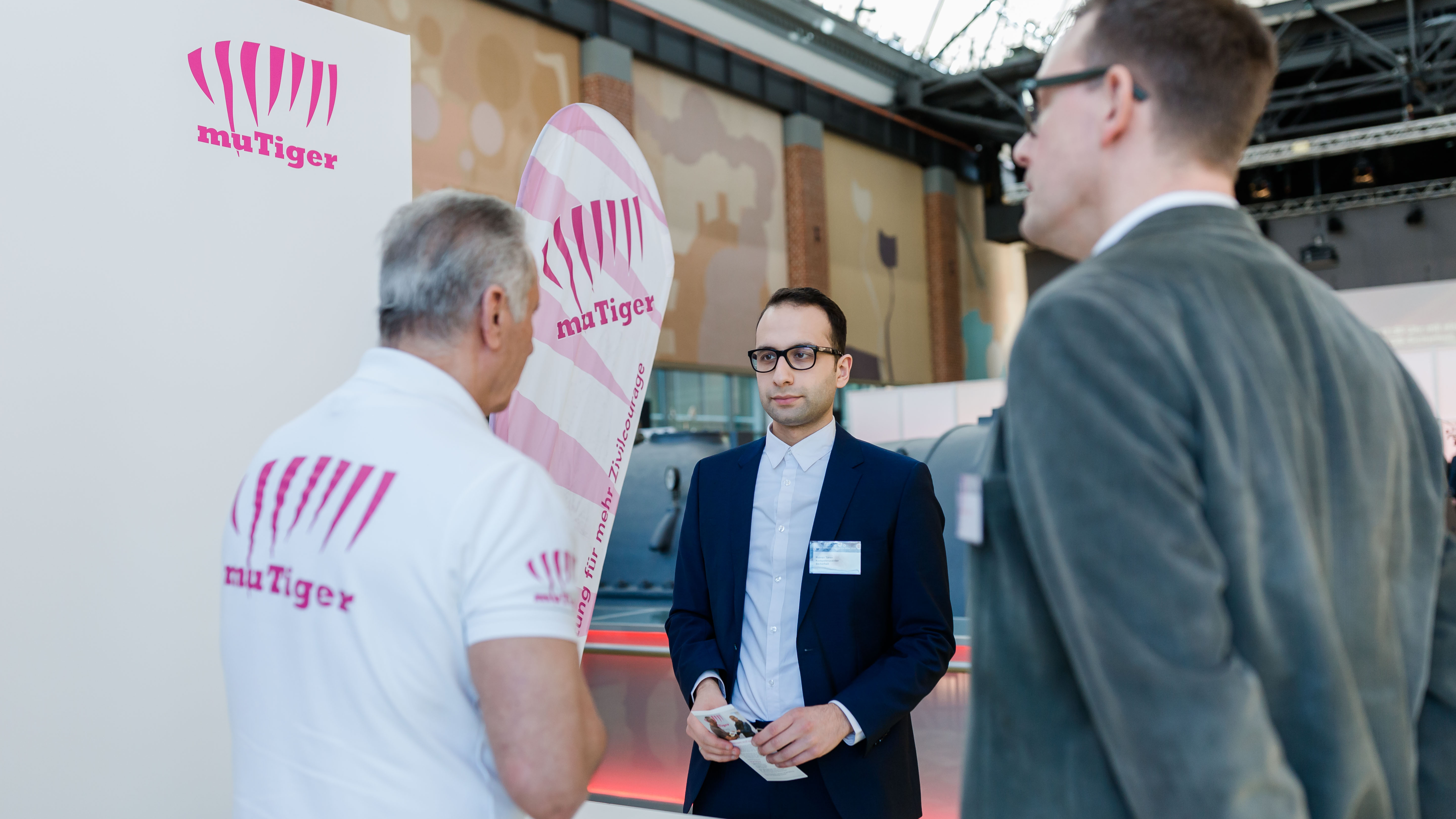 Ein Mitarbeiter von "muTiger" erklärt zwei Männern am Stand das Projekt. - öffnet Bild in größerer Ansicht